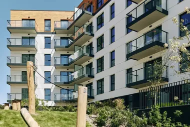 Nowoczesny apartamentowiec z balkonami i przeszklonymi balustradami, otoczony zielenią.